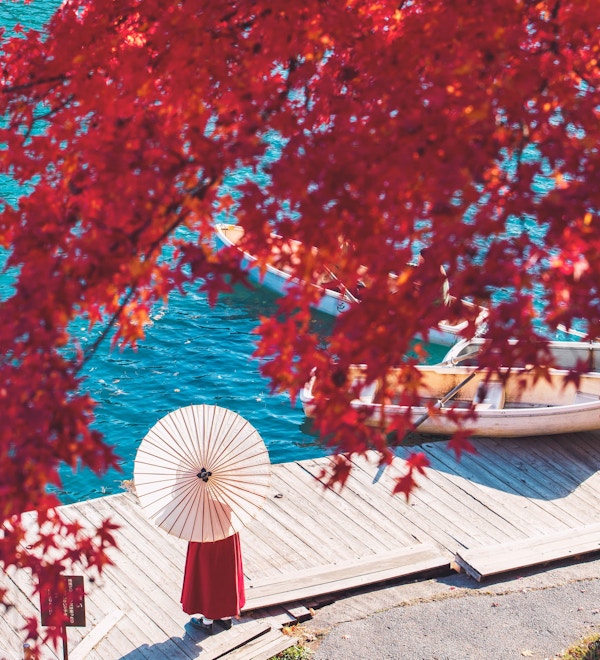 Kvinne med parasoll står ved blått vann og med rød japansk lønn i forkant