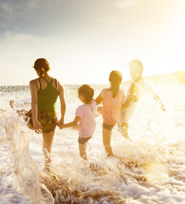 Familie med to barn fotografert bakfra mens de leker i vannet på en solrik strand.