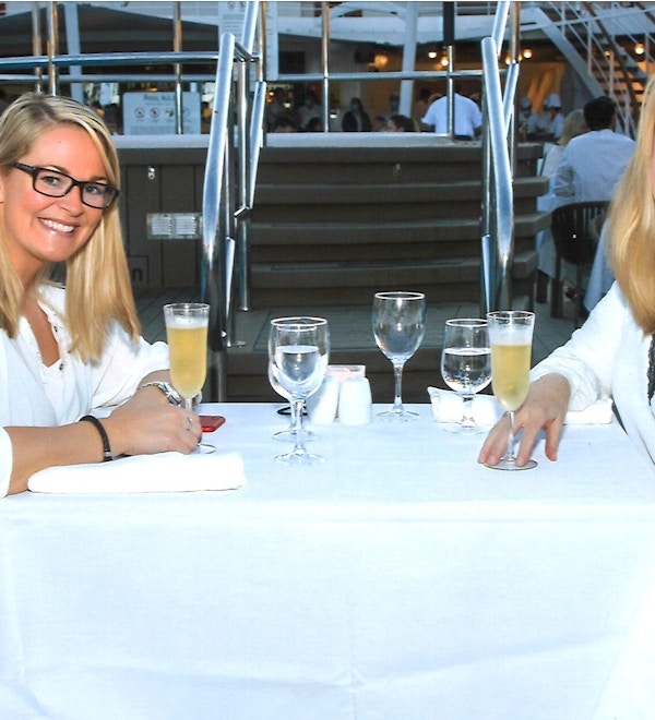 To damer drikker musserende vin, kledd i hvitt ved restaurantbord.