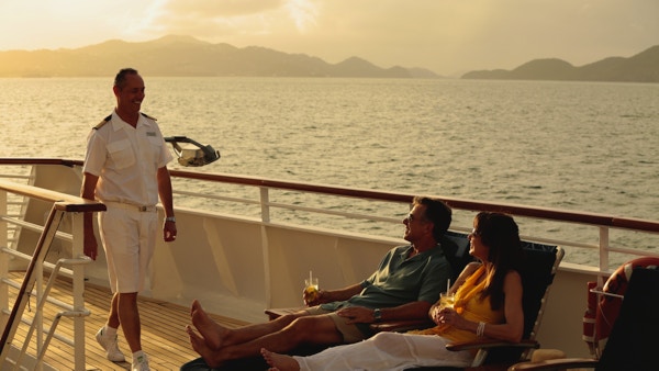 Par på dekk på yacht med drinker, en ansatt kommer smilende forbi.