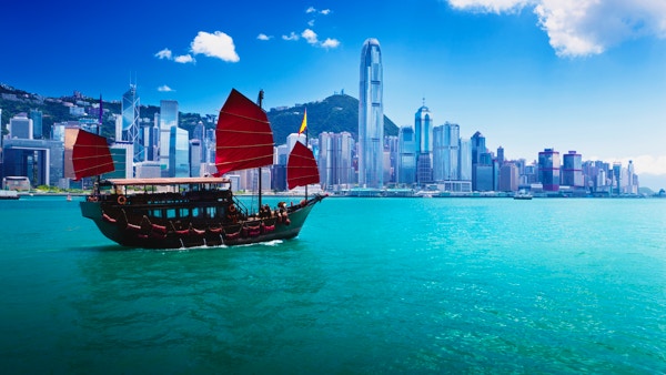Hongkongs skyline fra havnen på dagtid. Vannet er blågrønt, og et tradisjonelt kinesisk søppelskip med firkantede røde seil ligger i vannet i forgrunnen. Bygningene i horisonten er av ulik høyd og for det meste hvite foran en åsside dekket av mindre bygninger under en blå himmel.
