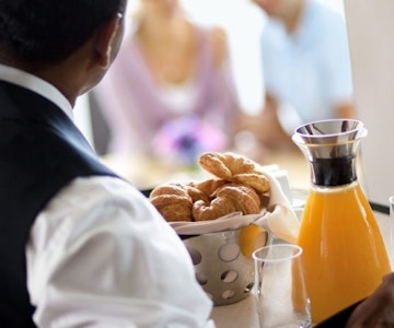 Kelner fotografert bakfra med brett med croissants og appelsinjuice med et par i bakgrunnen.