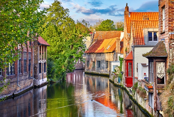Brugge, Belgia. Middelalderske gamle hus laget av gamle murstein ved vannkanal med båter i gamlebyen. Sommersolnedgang med solskinn og grønne trær. Pittoreske landskap.