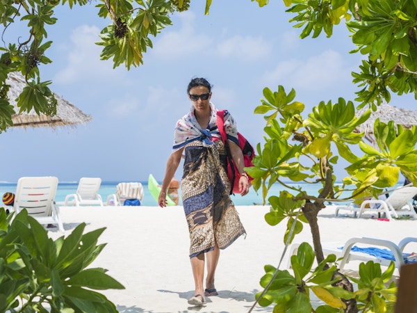Vindusvisning av vakker ung kvinne som bærer en sarong og går på en bikinistrand på Gulhi-øya, Maldivene. I bakgrunnen står solstoler med solbeskyttende parasoller, Det indiske hav og skyer på blå himmel ligger i bakgrunnen. Gulhi-øya ligger ikke langt fra Maafushi-øya på Maldivene, og er åpen for utlendinger.