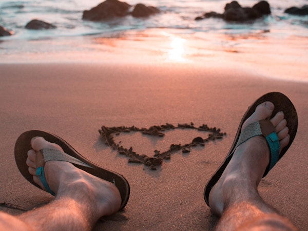 Mann i sandaler har risset inn et hjerte i sanden mens han ser på solnedgangen