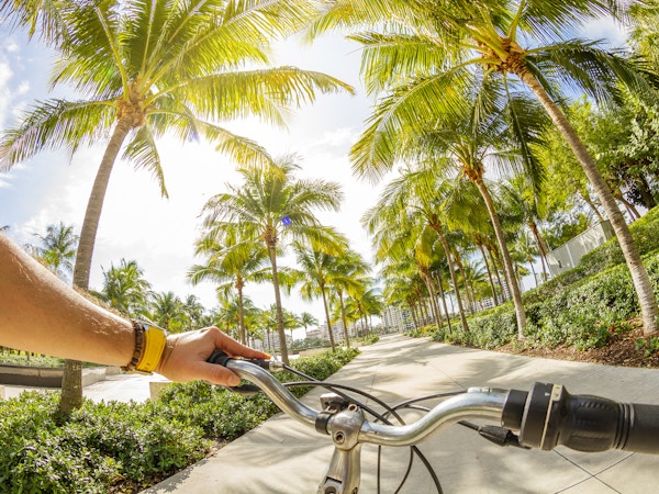 POV Synspunktbilde av en ung latinsk sportsmann som sykler barbeint en sykkel foran et turistisk Art Deco-distriktslandskap ved South Pointe Park i Miami Beach, Miami, Sør-Florida, USA - USA. På bakgrunn, det berømte og turistiske Art Deco-distriktet. Fotografering fra et personlig perspektiv i en eksotisk tropisk strandferie.