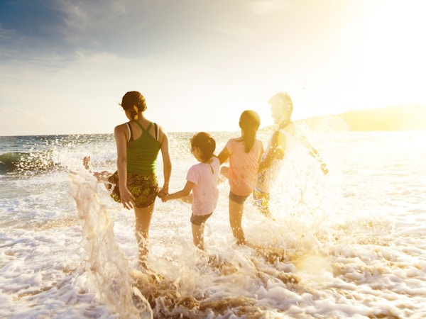 Familie med to barn fotografert bakfra mens de leker i vannet på en solrik strand.