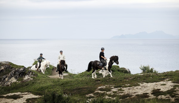 Tre personer rir på hest i kystlandskap.