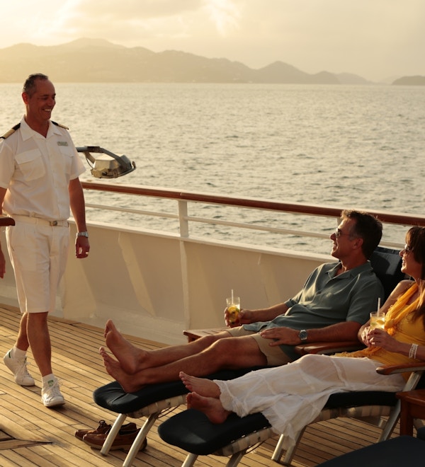 Par på dekk på yacht med drinker, en ansatt kommer smilende forbi.