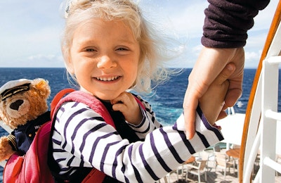 Jente holder hånda til en voksen på cruiseskip, har med sekk med bamse i.