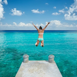 En kvinne hopper i blått vann fra bryggeutsikt fra baksiden på karibisk ferie