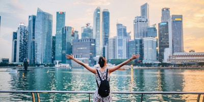 Ung reisende kvinne med armene utstrakt ser på solnedgangen over den fantastiske silhuetten i Singapore Bay-området. Frihet, reise, konsepter.